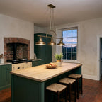 Quoizel Emery 3 Light Bar Pendant in Weathered Brass lit over a long oak-topped island in a green Victorian farmhouse kitchen